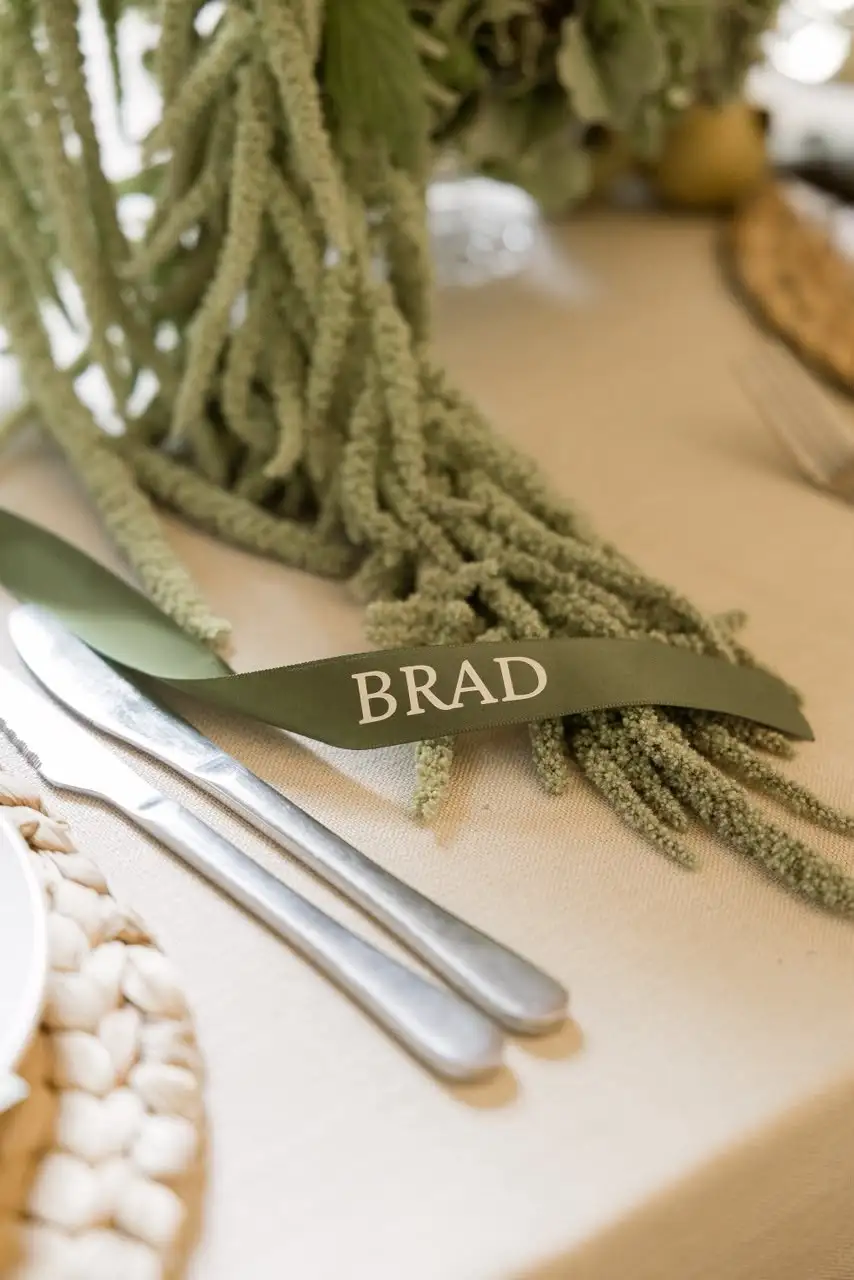 Olive green ribbon tag printed with a name, laid over cutlery and greenery