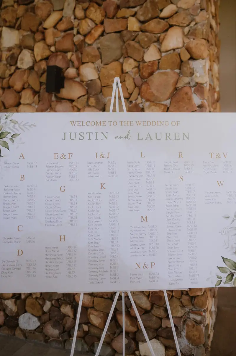Printed seating chart on a white easel against a stone wall, listing tables A to W