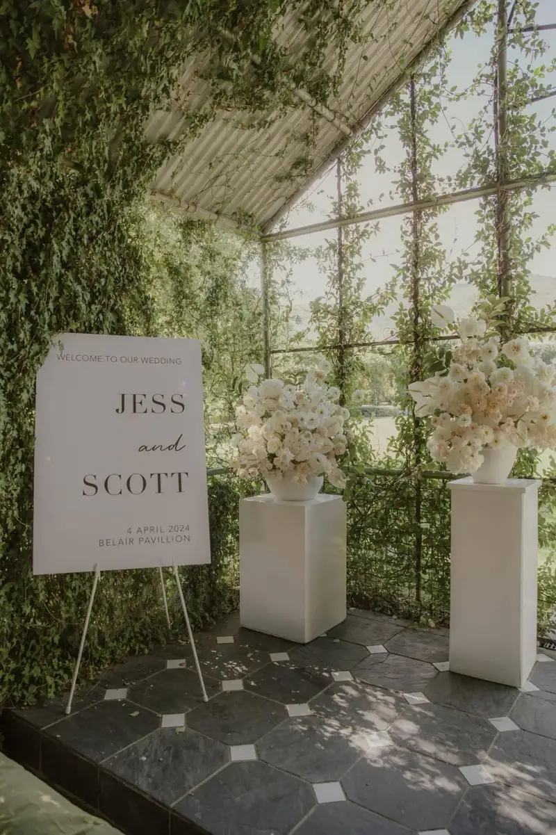 Welcome sign on an easel beside white florals under a vine-covered pergola